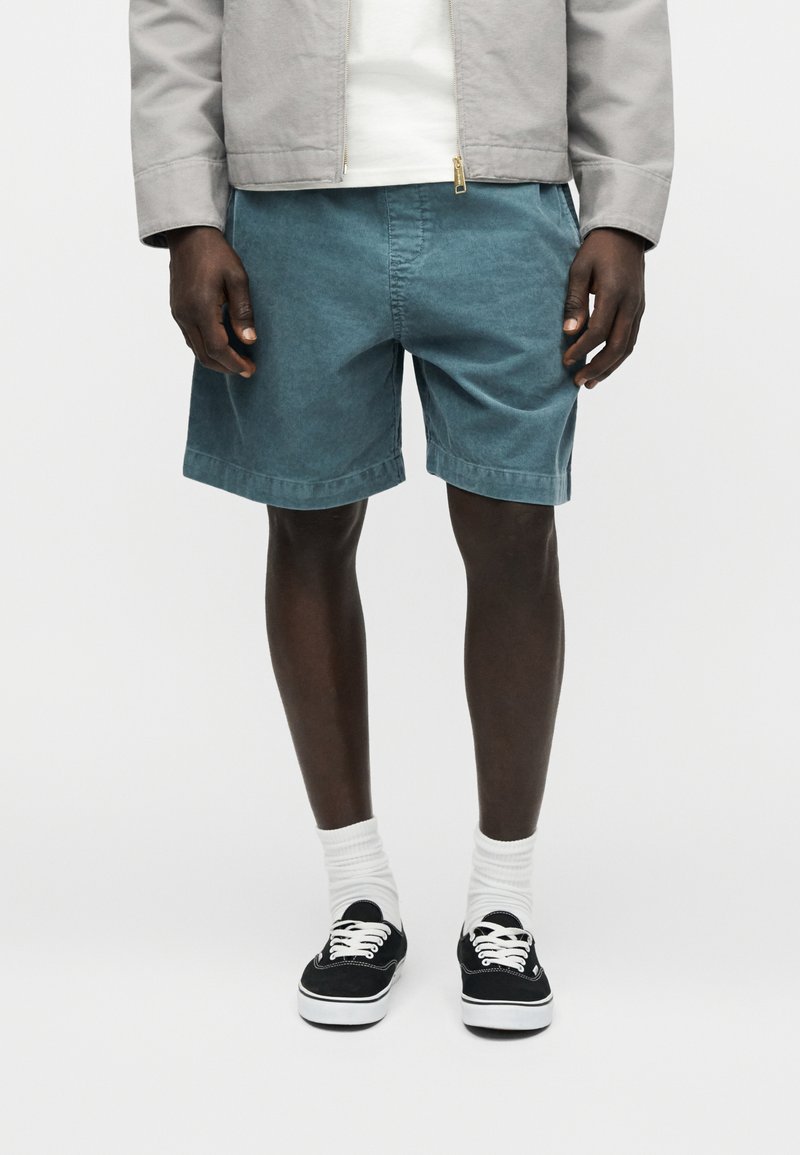 Homme portant un short en velours côtelé teal, des chaussettes blanches, des baskets noires, une chemise blanche et une veste gris clair, debout devant un fond uni.