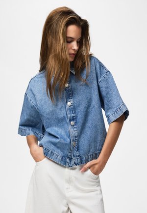 Ung kvinne med langt brunt hår iført en løs kortermet blå denimskjorte og hvite bukser, hendene i lommer, ser ned.