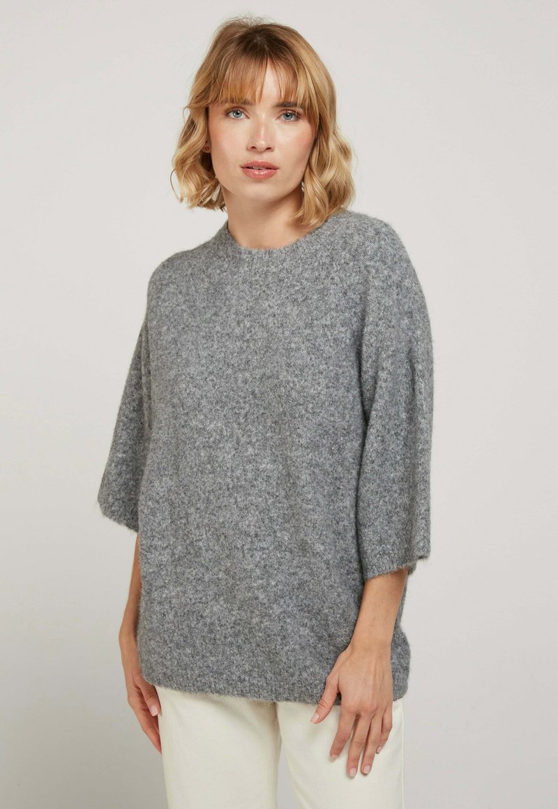 Pull en laine gris, flou, avec un col rond, des manches courtes et une coupe décontractée. Texture douce, design simple, sans motifs ni accessoires.