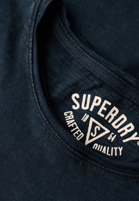 Camiseta de algodón azul marino con escote redondo. Presenta una etiqueta impresa con el logo "SUPERDRY" y "CRAFTED QUALITY" en texto blanco.
