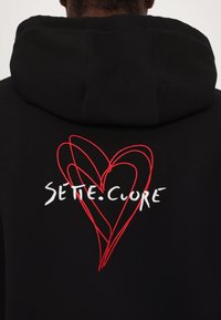 Haut à capuche noir avec cordon de serrage, présentant un contour de cœur brodé rouge et le texte blanc "SETTE.CUORE" au dos. Texture lisse.