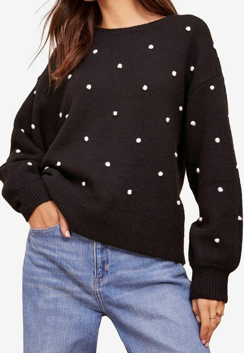 Femme portant un pull noir avec de petits pompons blancs et un jean bleu clair, la main posée dans la poche avant.