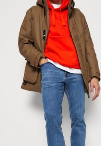Brown padded jacket over an orange hoodie, paired with blue jeans. Features zippers and pockets, with a contrasting white shirt underneath.