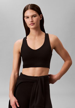 Młoda kobieta o długich brązowych włosach, ubrana w czarny biustonosz sportowy i czarne legginsy, stoi z jedną ręką na biodrze na szarym tle.