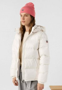 Witte gewatteerde jas met capuchon, voorzien van een ritssluiting aan de voorkant en zijzakken. Gecombineerd met een roze gebreide beanie en een beige trui.