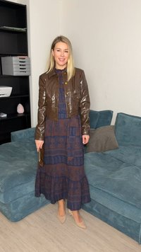 Une femme se tient à l'intérieur, portant une veste en cuir marron, une robe longue à motifs, des talons nus, et tenant une pochette marron près d'un canapé bleu avec des coussins.