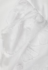 Tissu blanc avec section froncée et broderie florale texturée, montrant un tissage fin et des motifs délicats en relief sur une matière lisse.