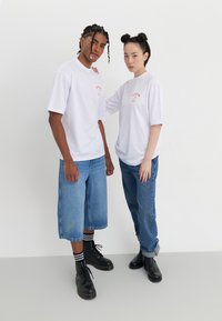 Baltos per dideles t-shirts, raudonas grafikos tekstas, derinamas su džinsiniais šortais ir džinsais. Modelis vilki juodus raištelius batus ir kulkšninius batus.