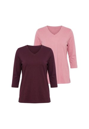Femme aux cheveux blonds ondulés portant un t-shirt violet foncé à col en V et un pantalon bleu, reposant une main sur la tête et l'autre dans la poche.