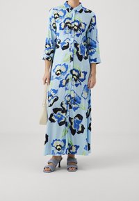 Maxi šaty z lehké modré látky s květinovým vzorem v modré, černé a zelené barvě. Mají zapínání na knoflíky vpředu a tříčtvrteční rukávy.