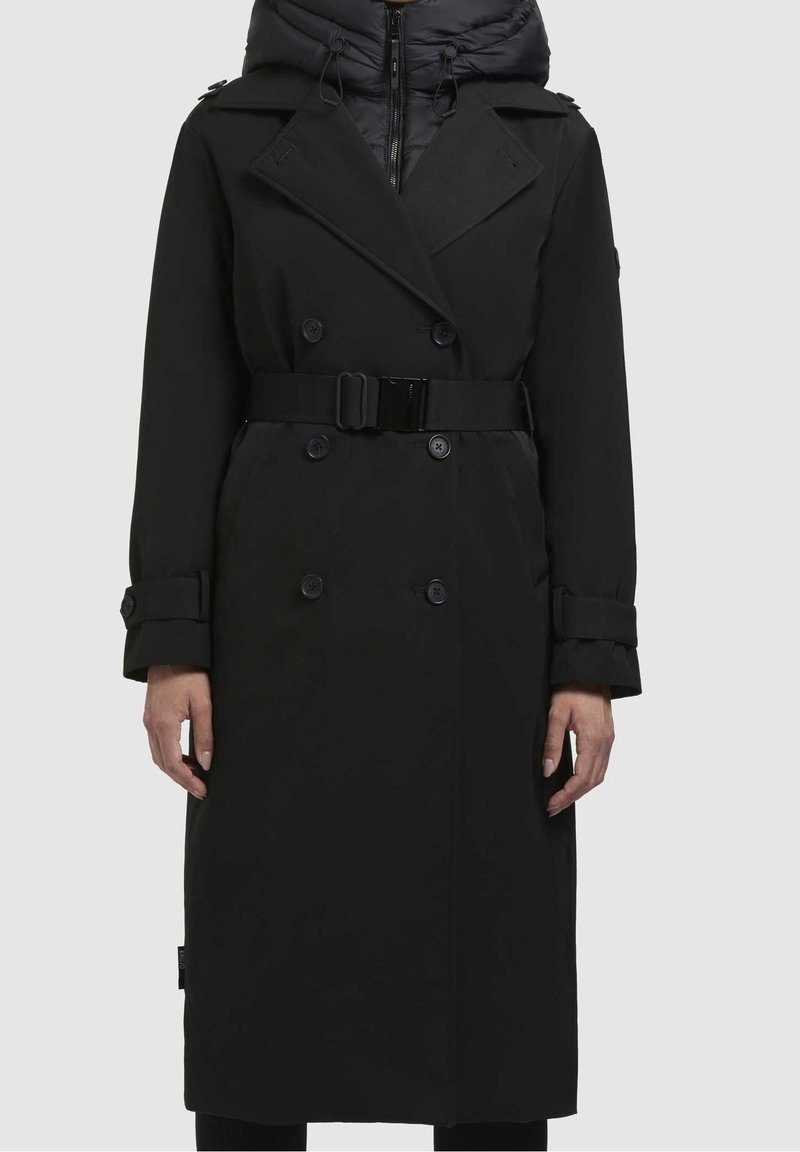 Long trench-coat noir ceinturé avec des boutons croisés porté sur une veste noire à capuche zippée.
