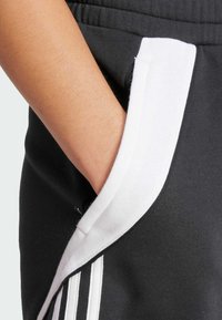 Pantaloni della tuta neri con rifinitura bianca sulle tasche e tre strisce bianche lungo il lato. Il tessuto sembra morbido e texturizzato.