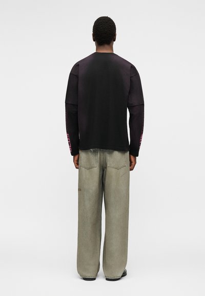 Άνδρας με κοντά σγουρά μαλλιά που φοράει μαύρο μακρυμάνικο πουκάμισο με ροζ γράμματα στα μανίκια και φαρδιά ανοιχτόγριζα τζιν, στέκεται με την πλάτη γυρισμένη.