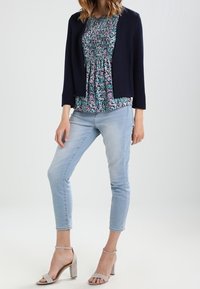 Žena ve tmavě modré blazer, květinové blouse, světle modrých zkrácených džínách a béžových sandálech na blokovém podpatku s kotníkovým páskem, stojící s jednou nohou pokrčenou.