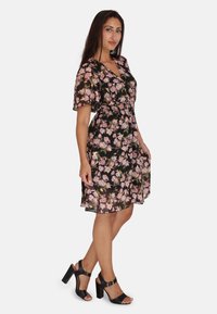 Vestido envolvente floral de tejido negro transparente con patrones en rosa y verde. Cuenta con mangas cortas, una cintura fruncida y fluye hasta la mitad del muslo.