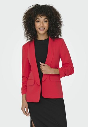 Vrouw met krullend haar die een rood colbert draagt over een zwarte jurk met een zijsplit, glimlachend en staand tegen een effen achtergrond.