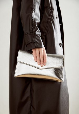 Hånd i sort læderjakke, der holder en skinnende sølvkuvertclutch mod en neutral baggrund.