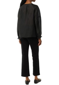 Schwarze, semi-transparente Bluse mit langen Ärmeln und gerafften Manschetten, kombiniert mit schwarzen, taillierten Hosen und flachen Schuhen. Schlichtes, minimalistisches Design.