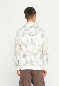 Felpa chiara con un motivo a foglie grigio. Presenta un cappuccio con cordini e orlo a costine. Abbinata a pantaloni cargo marroni.