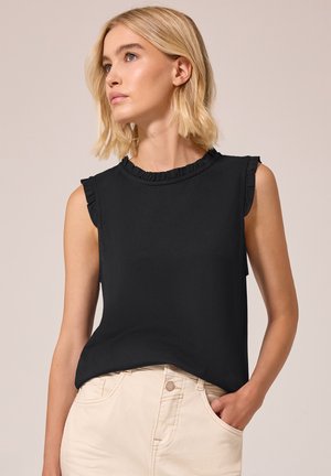 Femme blonde portant un haut noir sans manches à bords froncés et un pantalon beige, regardant sur le côté avec une main dans la poche.