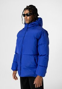 Blauwe pufferjack met een capuchon, voorzien van een ritssluiting aan de voorkant, brede mouwen en een doorgestikte ontwerp. Model draagt zwarte zonnebril.