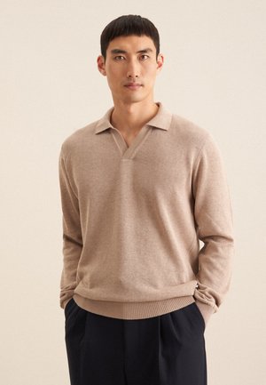 Beige gebreide pullover met een V-hals en korte kraag, voorzien van geribbelde manchetten en onderzoom, gedragen over donkere broeken, tegen een neutrale achtergrond.