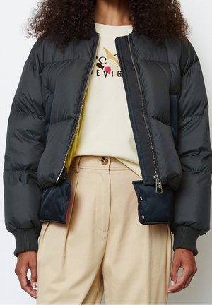 Vrouw draagt een zwart donsjack over een crèmekleurige trui met print, beige hoge taille broek, staand tegen een effen achtergrond.