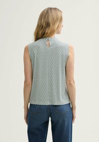 Bezramenný top v bielej farbe s námorníckym geometrickým vzorom, s detailom otvoru na chrbte. Kombinovaný s tmavomodrými džínovými nohavicami.