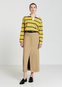 Gelb- und grün gestreiftes Langarm-Polohemd mit einem weißen Kragen, kombiniert mit einem beigen langen Rock mit Schlitz vorne und einem schwarzen Gürtel. Schwarze Slipper.