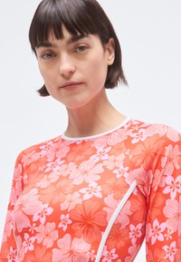 Červený dlouhý rukáv rash guard s velkými vzory růžových a bílých květin, z lehkého materiálu a s bílým límečkem jako akcentem.