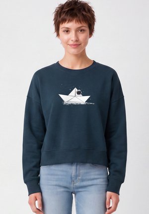 Femme portant un sweat-shirt bleu foncé avec un motif de bateau en papier blanc et d'astronaute, associé à un jean bleu clair, debout devant un fond blanc.