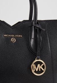 Sac à main en cuir texturé noir Michael Kors avec plaque de logo dorée et pendentif rond doré MK suspendu à la poignée.