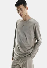 Långärmad sporttröja i ljusbeige, tillverkad av fuktavvisande tyg. Har en rund halsringning och en liten logotyp på bröstet.