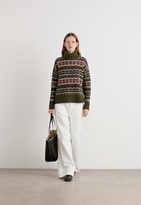 Gehaakte coltrui in olijfgroen met meerkleurige geometrische patronen; gedragen met witte wijde broek en een zwarte handtas.