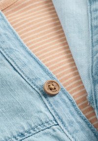 Hellblaues Denim-Jackett, das mit einem beige-weißen gestreiften Hemd darunter präsentiert wird, mit einem Holzknopfdetail an der Jacke.