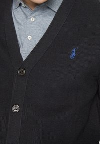 Svart stickad cardigan med v-ringning, med en blå polo-logotyp på vänster bröst, knappstängning, bärs över en ljusgrå skjorta med krage.