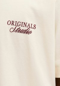 Μπεζ μπλούζα με κεντημένο σε μπορντό κείμενο "ORIGINALS Studio." Μαλακό βαμβακερό υλικό με λεία υφή και στρογγυλή λαιμόκοψη.