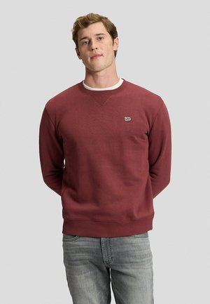 Hombre de pie frente a un fondo liso, con sudadera burdeos de Lee y vaqueros azul claro, con las manos detrás de la espalda y expresión facial neutra.