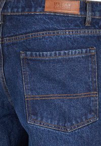 Tummansininen farkkukangas, jossa on oranssia tikkausta, takatasku ja ruskea nahkaleima, jossa lukee "Urban Classics Denim Factory."