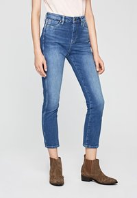 Högmidjade blå denimjeans med slim fit, kort längd, lätt slitning och klassisk femfickdesign, tillsammans med bruna ankelstövlar.