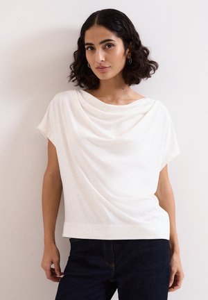 Femme aux cheveux courts, bouclés et foncés, portant un haut blanc drapé à manches courtes et un pantalon foncé, debout contre un mur clair.