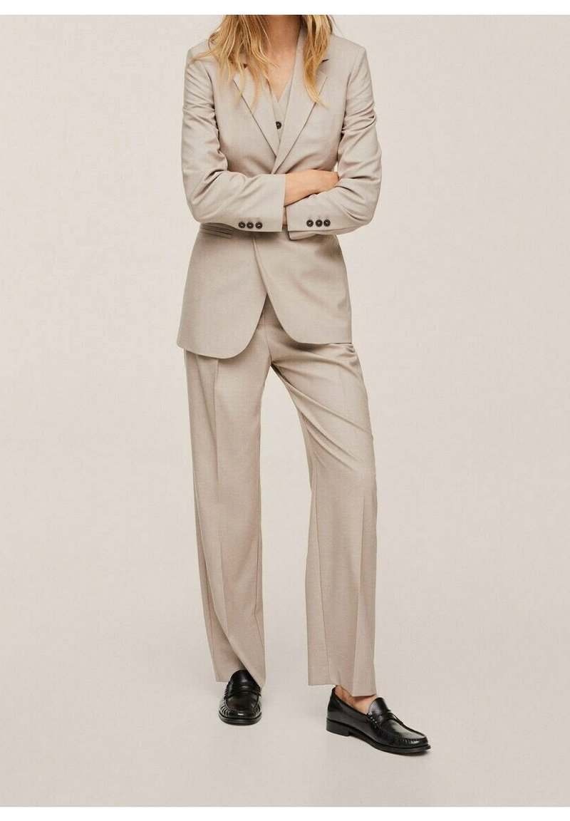 Beżowy garnitur na miarę z marynarką z klapami z wycięciem i szerokimi spodniami. Gładka faktura, jednorzędowe zapięcie na jeden guzik, w połączeniu z czarnymi mokasynami.