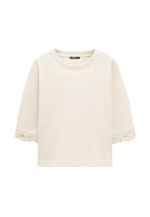 Kremowy top z krótkim rękawem o okrągłym dekolcie, z koronkowym wykończeniem na końcach rękawów, prezentowany na białym tle.