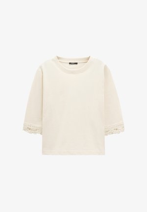 Kremowy top z krótkim rękawem o okrągłym dekolcie, z koronkowym wykończeniem na końcach rękawów, prezentowany na białym tle.