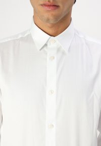 Camisa blanca de botones confeccionada con tela suave. Presenta un cuello clásico, cierre frontal con botones y un diseño liso sin patrones visibles.