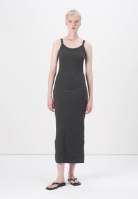 Długa szara dzianinowa sukienka o dopasowanym kroju, z cienkimi ramiączkami i teksturowanym wzorem. Noszona z czarnymi płaskimi sandałami. Minimalistyczny design na białym tle.