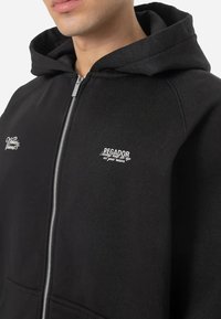 Crna zip majica napravljena od mješavine pamuka, s kapuljačom, prednjim džepovima i bijelim izvezenim logotipima na lijevoj strani prsa i rukavu.