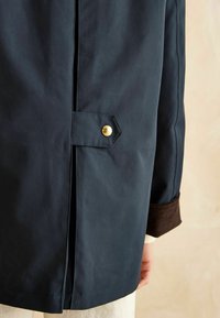 Palton impermeabil de culoare bleumarin, cu o închidere vizibilă cu capse aurii, fante laterale și manșete din corduroy maro, evidențiind o textură de țesătură mată și netedă.