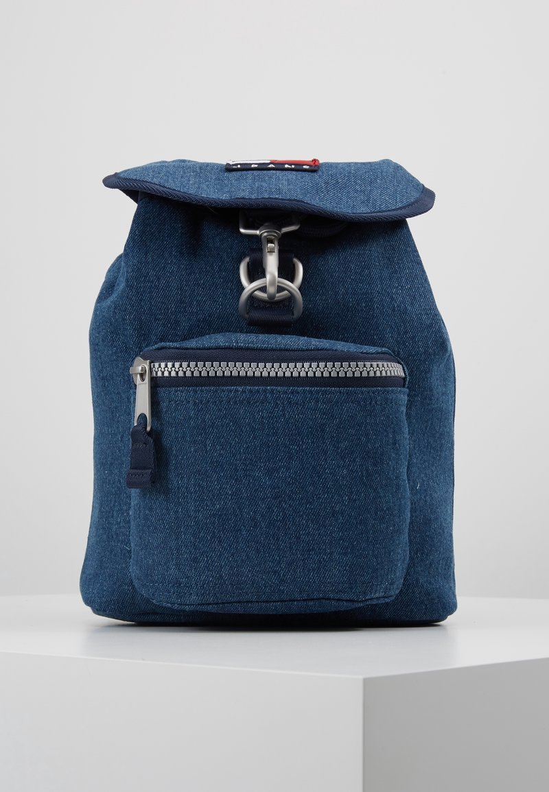 Petit sac à dos en denim bleu avec poche zippée à l'avant et fermeture à boucle métallique, posé sur une surface blanche sur un fond uni.