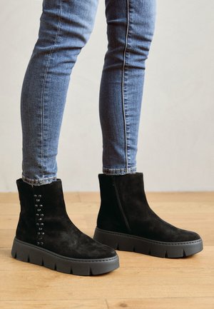Black suede ankle boots with a chunky gray sole, side zipper, and decorative black studs along the outer ankle. Textured surface and shape.